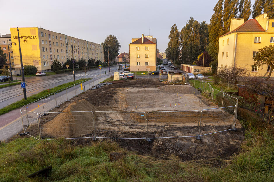 na zdjęciu ogrodzony plac budowy, widać rozkopaną działkę, w tle trzy bloki mieszkalne