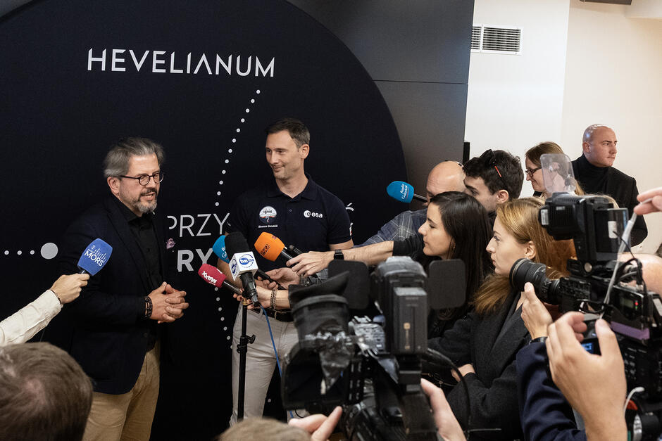 Dwóch mężczyzn przy mikrofonach otoczeni tłumem dziennikarzy.