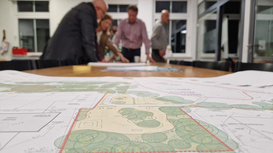 Na pierwszym planie duży wydruk z koncepcją zagospodarowania, na drugim planie ludzie pochyleni nad planami