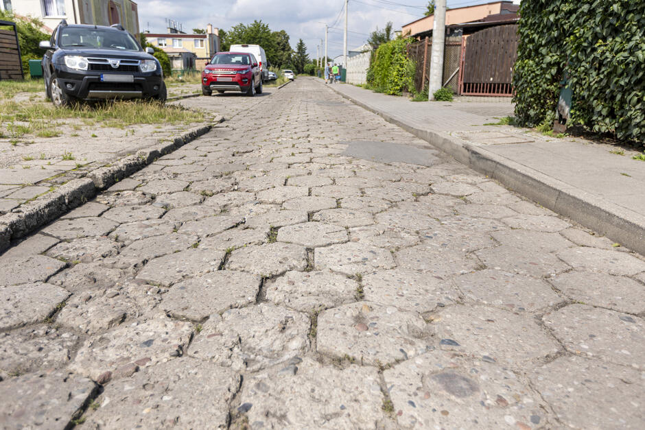 na zdjęciu nawierzchnia ulicy wykonana z betonowych płytek, tak zwanej trylinki, w tle widać kilka stojących samochodów