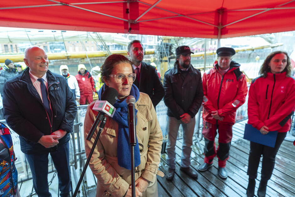 Kobieta w beżowym płaszczu i niebieskim szaliku przemawia do mikrofonu podczas wydarzenia pod czerwonym namiotem. Za nią stoją przedstawiciele organizatorów i uczestnicy spotkania, a w tle widać fragment żaglowca przy nabrzeżu.
