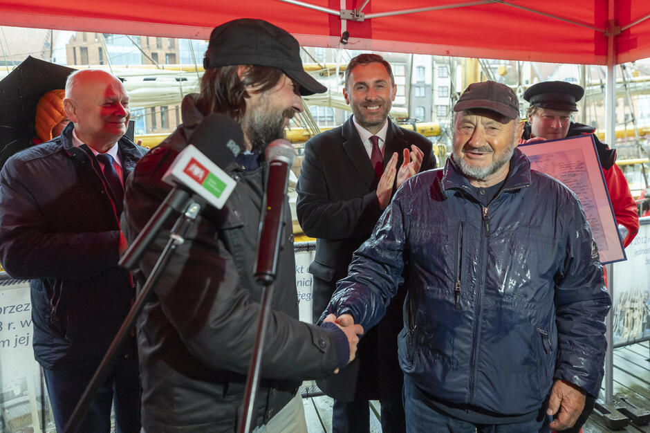 Pod zadaszeniem na nabrzeżu mężczyźni wymieniają uścisk dłoni podczas oficjalnego spotkania, a obok stoją osoby bijące brawo. W tle widać żaglowiec i elementy portowego otoczenia Gdańska.
