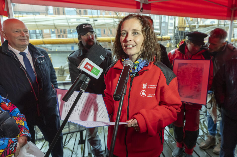 Kobieta w czerwonej kurtce z logo Uniwersytetu Wrocławskiego i ekspedycji GeoArctic przemawia do mikrofonu, trzymając w ręku kartkę z tekstem. Za nią stoją uczestnicy wydarzenia oraz żaglowiec zacumowany przy nabrzeżu.

