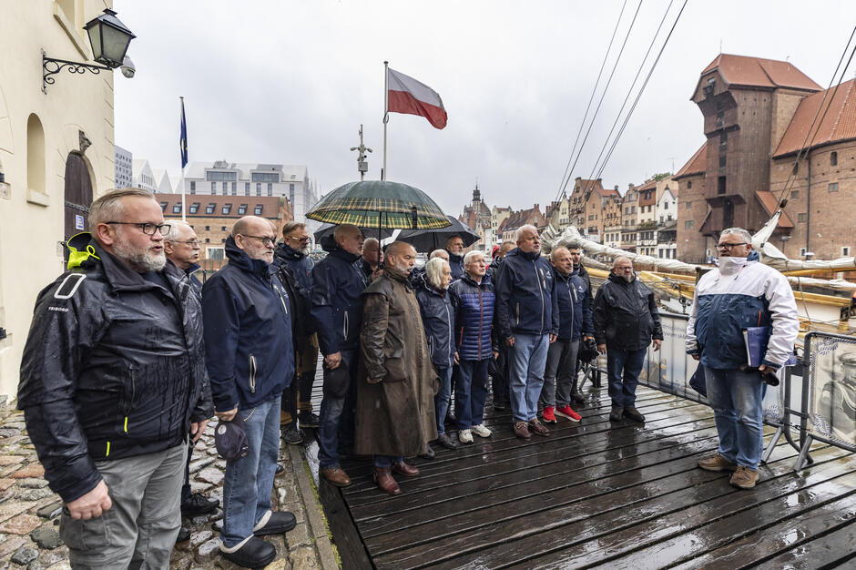 Grupa osób stoi na nabrzeżu w Gdańsku podczas deszczowej pogody, słuchając mężczyzny przemawiającego z teczką w ręku. W tle widać żaglowiec, polską flagę oraz panoramę zabytkowych kamienic nad Motławą.
