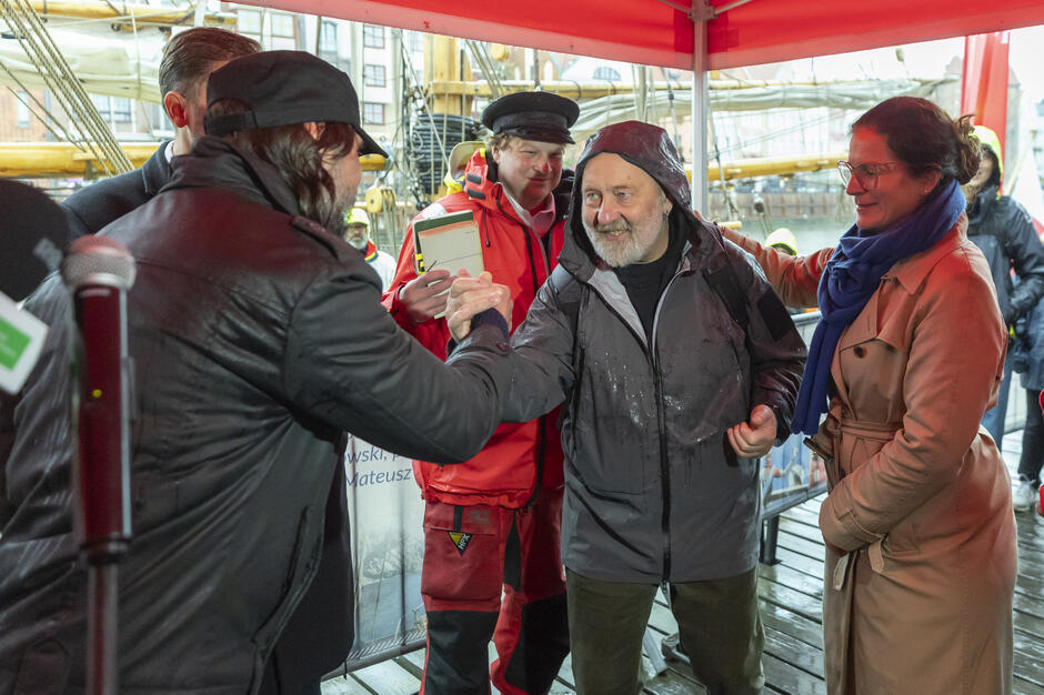 Pod czerwonym namiotem dwaj mężczyźni serdecznie ściskają sobie dłonie, a obok nich stoją uczestnicy wydarzenia w żeglarskich i przeciwdeszczowych strojach. W tle widoczny jest fragment żaglowca zacumowanego przy nabrzeżu w Gdańsku.
