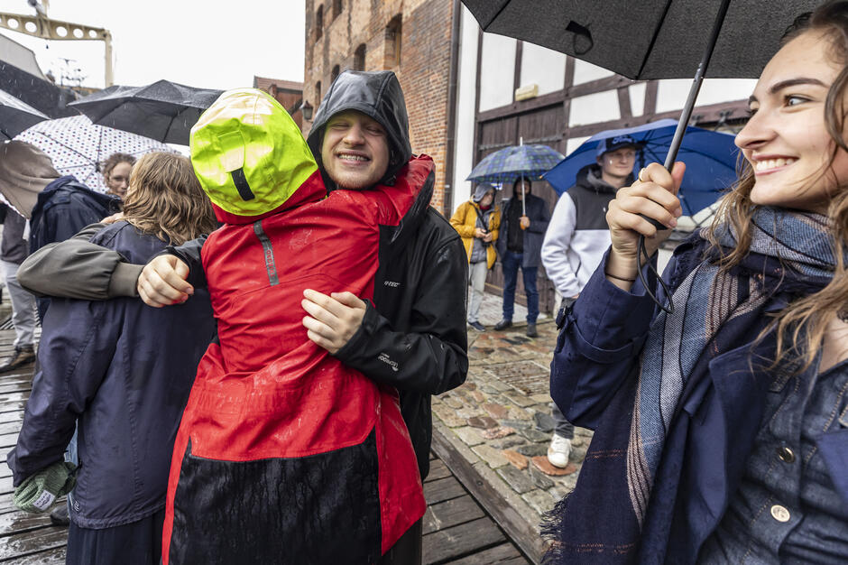 Młodzi uczestnicy wydarzenia witają się serdecznie w deszczu, przytulając się z radością mimo niesprzyjającej pogody. W tle widać osoby z parasolami oraz zabytkowe budynki nabrzeża w Gdańsku.
