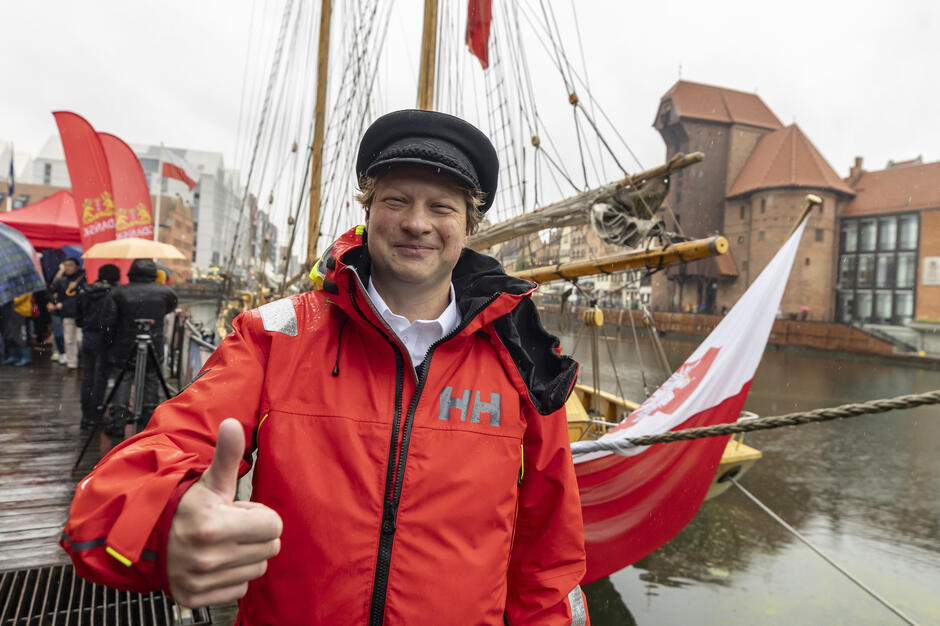 Mężczyzna w czerwonej kurtce żeglarskiej i czapce marynarskiej pokazuje gest „kciuk w górę”, stojąc przy nabrzeżu w deszczowy dzień. W tle widać żaglowiec z biało-czerwoną flagą oraz zabytkowe budynki Gdańska nad Motławą.

