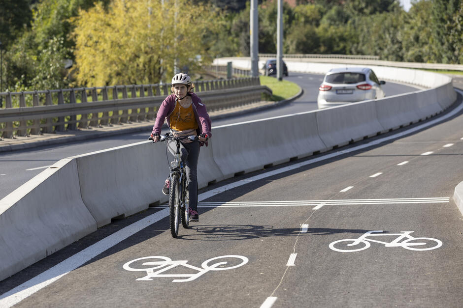 na zdjęciu kobieta jadąca rowerem, w kasku na głowie, jedzie oznakowaną drogą rowerową, w tle po lewej jadą dwa samochody jezdnią