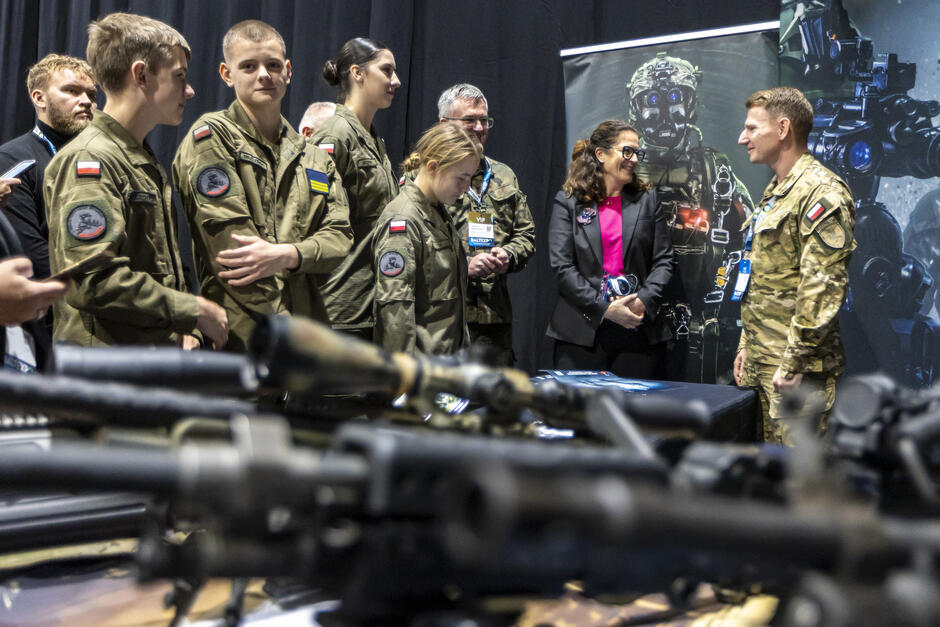 Kobieta w garsonce rozmawia z mężczyzną w mundurze w hali targów, dookoła młodzież w mundurach.