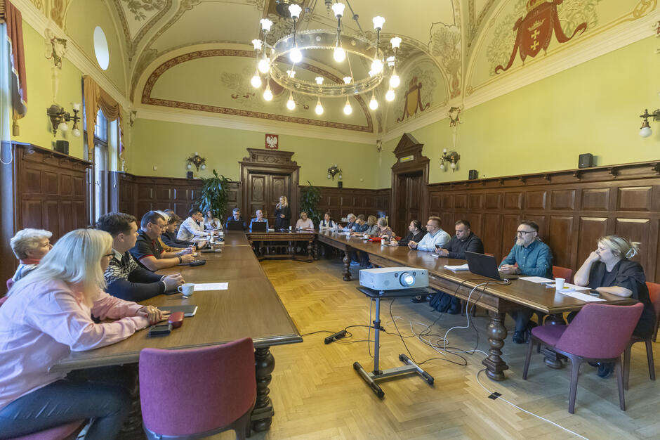 zabytkowa sala w której przy wielki stole w kształcie litery u siedzi kilkadziesiąt osób