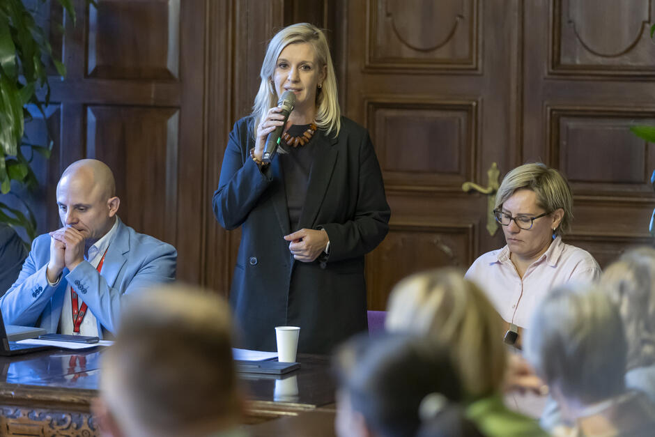 na zdjęciu kobieta w średnim wieku, w ciemnej marynarce, mówi do mikrofonu, stojąc za stołem