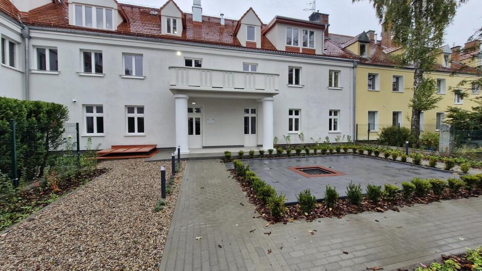 Biały budynek z balkonem podpartym dwiema kolumnami. Przed nimi placyk z trampoliną wpuszczoną w podłoże