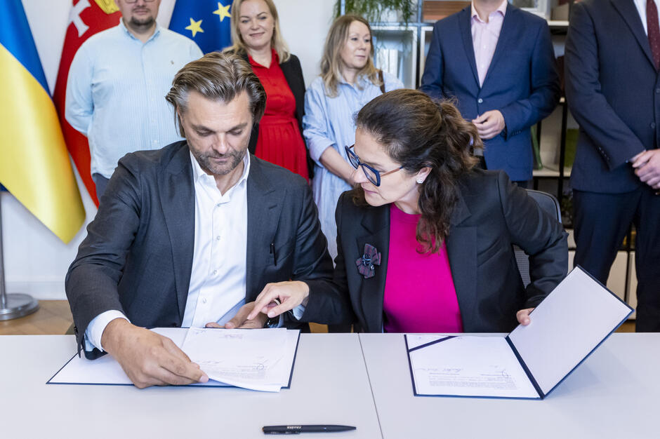 przy dużym stole siedzą dwie dorosłe osoby, podpisują umowę, za nimi stoi kilka osób w rzędzie