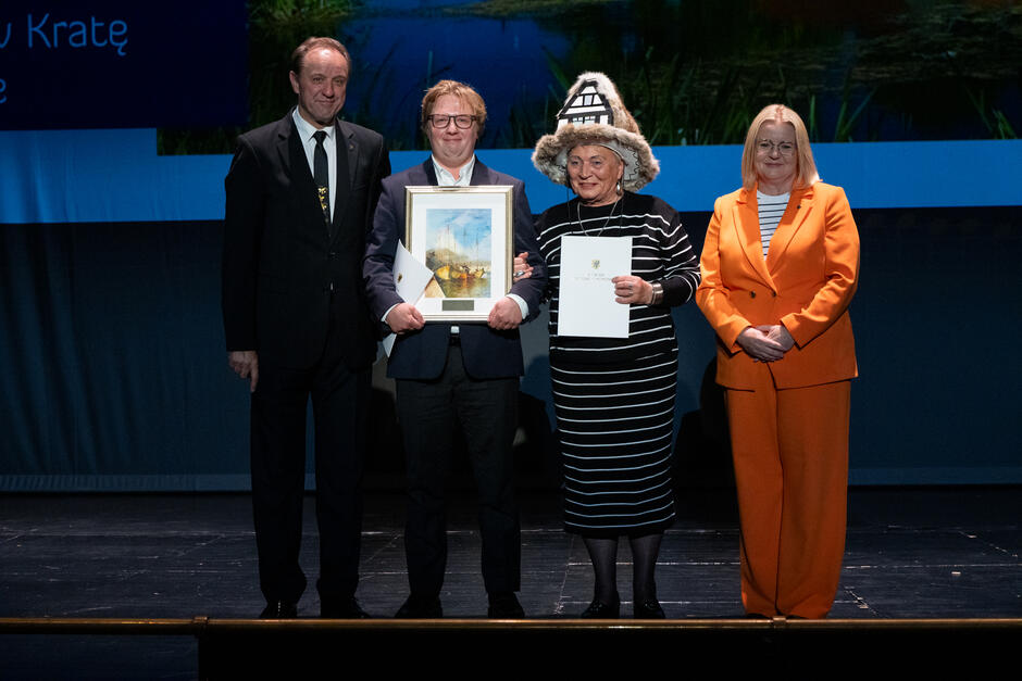 Cztery osoby stoją na scenie podczas uroczystego wręczenia nagród, trzymając dyplomy i oprawione wyróżnienie. Jedna z kobiet ma na sobie charakterystyczny kapelusz stylizowany na architekturę domów szachulcowych, co dodaje wydarzeniu regionalnego charakteru. 