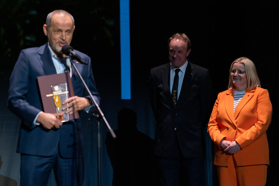 Mężczyzna w garniturze przemawia do mikrofonu, trzymając statuetkę i dyplom. Obok niego stoją dwie osoby obserwujące wystąpienie, w tym kobieta w pomarańczowym garniturze i mężczyzna w ciemnym garniturze. 