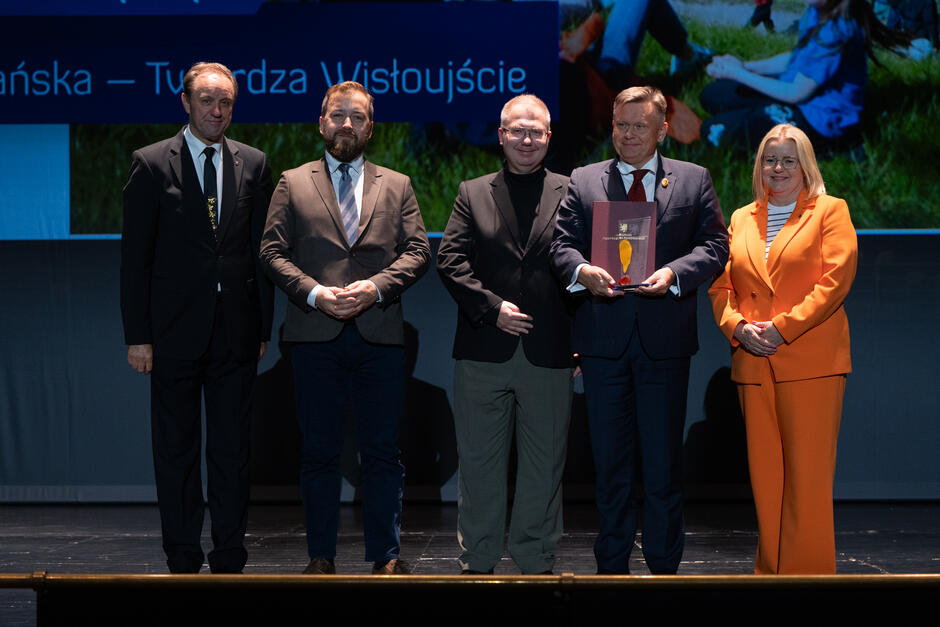 Pięć osób pozuje wspólnie na scenie podczas ceremonii wręczenia nagród, trzymając statuetkę i dyplom. W tle widoczny jest ekran z napisem informującym o wyróżnieniu dla Muzeum Gdańska – Twierdza Wisłoujście. 