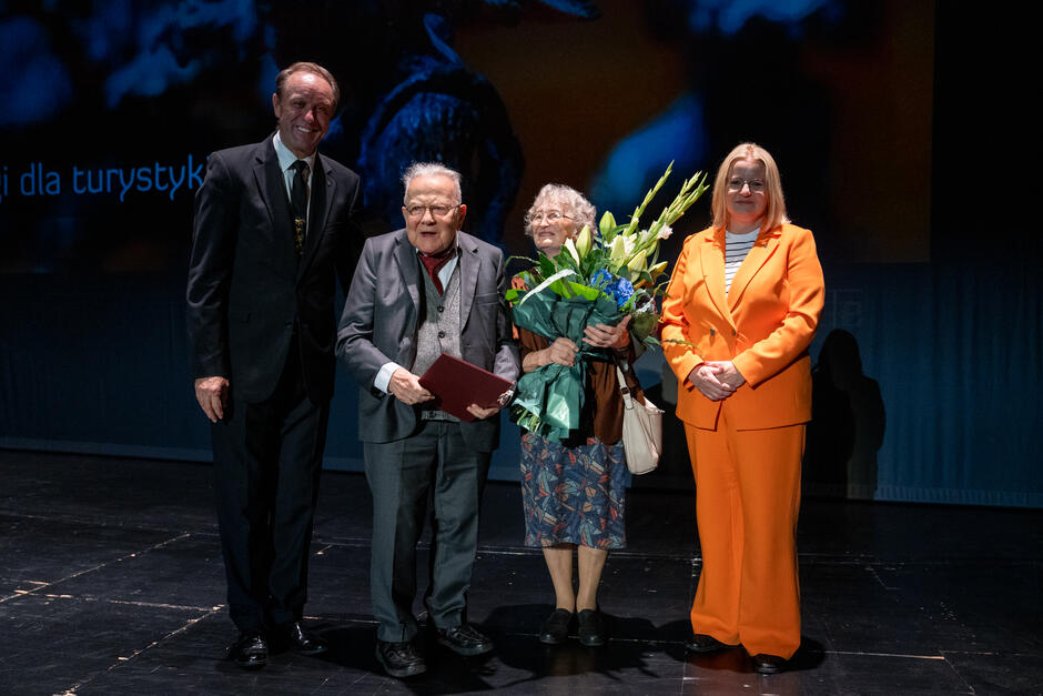 Cztery osoby stoją razem na scenie podczas uroczystości wręczenia nagród, w tym starsze małżeństwo z bukietem kwiatów i dyplomem. Obok nich stoją organizatorzy wydarzenia, uśmiechnięci i gratulujący wyróżnionym. 