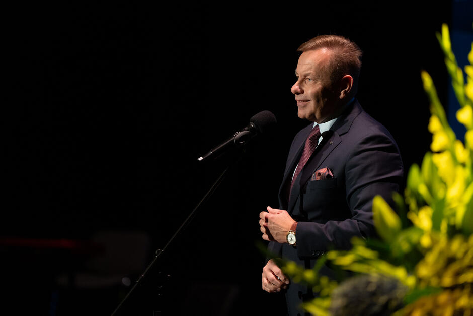 Mężczyzna w garniturze przemawia na scenie, stojąc przy mikrofonie. Na pierwszym planie widać żółte kwiaty, które dodają kadrowi uroczystego charakteru. 