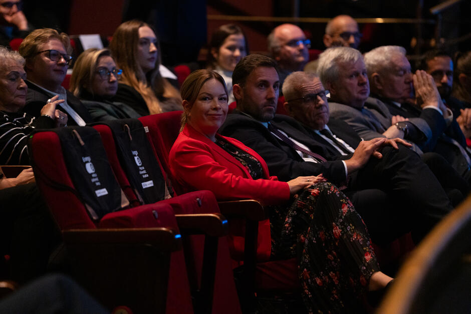 Uczestnicy wydarzenia siedzą na widowni, uważnie śledząc przebieg uroczystości. W centralnej części kadru kobieta w czerwonym żakiecie i mężczyzna w garniturze patrzą w stronę obiektywu, tworząc życzliwy i elegancki nastrój zdjęcia. 