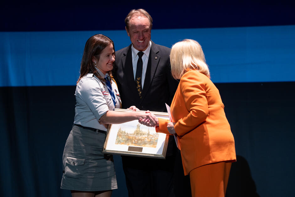 Kobieta w mundurze harcerskim odbiera nagrodę w formie oprawionego obrazu, ściskając dłoń kobiety w pomarańczowym garniturze. Za nimi stoi mężczyzna w garniturze, z uśmiechem obserwujący moment wręczenia wyróżnienia.
