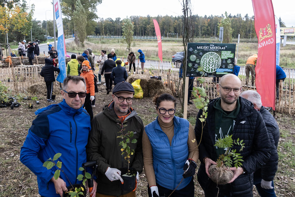 Trzech mężczyzn i kobieta pozują do zdjęcia z sadzonkami drzew w rękach. Za nimi grupa ludzi sadzi drzewka.