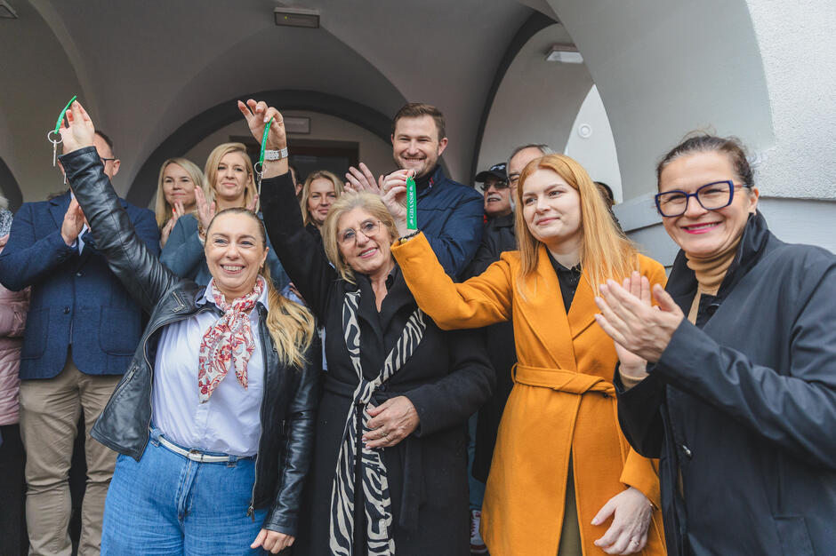 Grupa osób śmieje się i pozuje do zdjęcia przed wejściem do budynku. Kilka z nich pokazuje klucze do budynku.