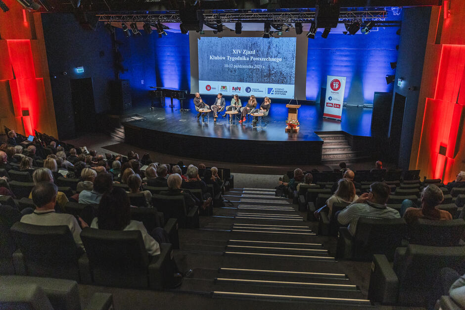 W sali kilka osób siedzi na fotelach na scenie, na publiczności siedzą słuchacze.