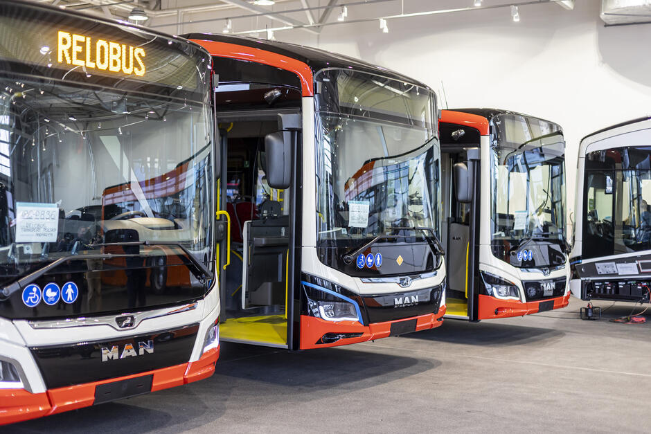 Na zdjęciu widać nowoczesne autobusy miejskie marki MAN stojące w hali produkcyjnej lub salonie wystawowym. Pojazdy mają czarno-biało-czerwone malowanie oraz wyświetlacze z napisem „RELOBUS”. Wnętrza autobusów są widoczne przez szerokie przednie szyby i otwarte drzwi. Na przednich częściach pojazdów umieszczone są piktogramy informujące o udogodnieniach dla osób z niepełnosprawnościami, wózków dziecięcych i pasażerów starszych. Cała scena oświetlona jest światłem sztucznym, podkreślającym nowoczesny charakter miejsca i pojazdów.