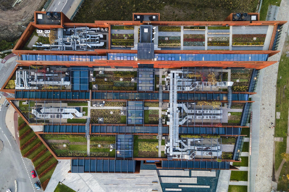 Zdjęcie z dronu, widok na dach budynku, na którym rośnie trawa i rośliny oraz są panele słoneczne i różne instalacje.