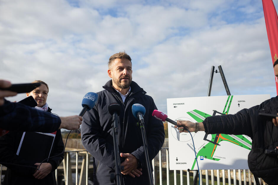 Mężczyzna udziela wypowiedzi mediom podczas konferencji prasowej na świeżym powietrzu, stojąc przed mapą przedstawiającą planowany układ drogowy. Obok niego stoi kobieta z dokumentami, a kilka mikrofonów skierowanych w jego stronę sugeruje obecność licznych dziennikarzy. 