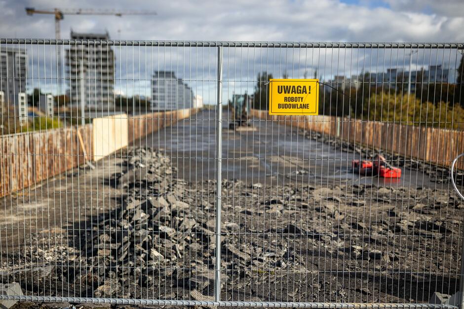 Zamknięty dla ruchu most jest w trakcie prac rozbiórkowych, o czym informuje żółta tabliczka z napisem „Uwaga! Roboty budowlane”. Na powierzchni widać stosy pokruszonego asfaltu i sprzęt budowlany w tle. 