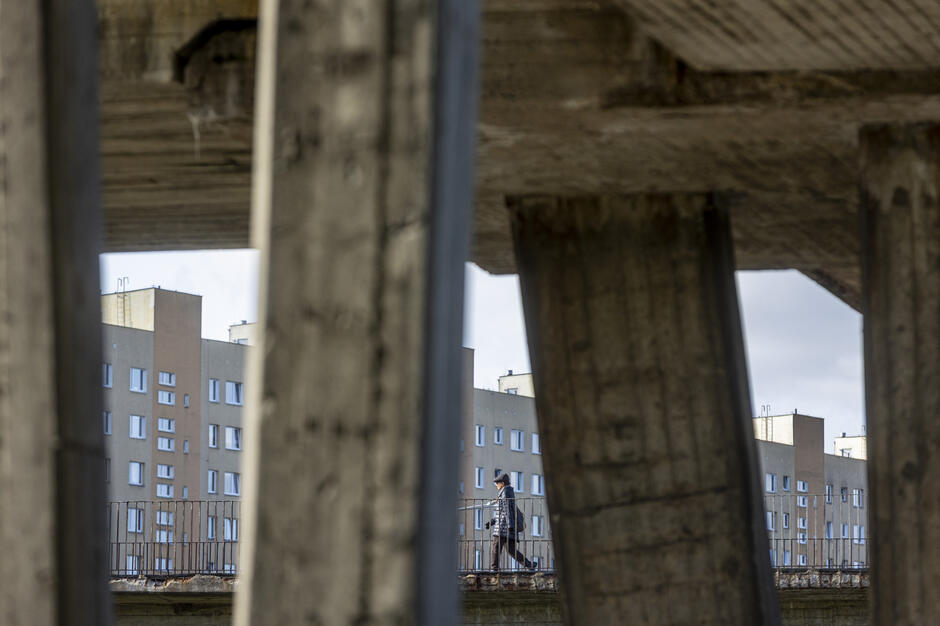 Przez betonowe filary wiaduktu widać przechodnia idącego po kładce dla pieszych. W tle znajdują się wysokie bloki mieszkalne, tworzące charakterystyczny miejski krajobraz. 