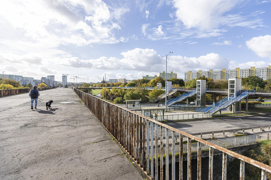 Osoba spaceruje z psem po szerokiej, zniszczonej kładce z widocznymi śladami korozji na barierkach. W tle widać osiedle bloków, niebieskie schody prowadzące do wind oraz rozwijającą się infrastrukturę miejską otoczoną zielenią.
