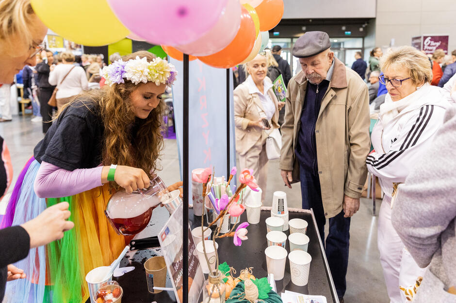 Grupa ludzi zebranych wokół stołu z różnymi przedmiotami na nim. stół jest ozdobiony kolorowymi balonami i kwiatami. po lewej stronie młoda dziewczyna w tęczowej sukience i koronie kwiatowej na głowie. wlewa drinka do szklanki i wydaje się wchodzić w interakcje z ludźmi wokół niej. Obok niej starszy mężczyzna w beżowym płaszczu i kapeluszu z zainteresowaniem patrzy na dziewczynę. W tle można zobaczyć inne osoby spacerujące i przeglądające sklep.