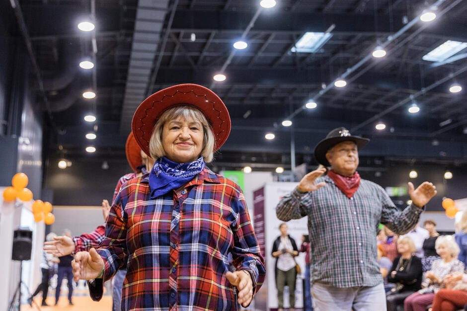Kobieta w czerwonym kapeluszu kowbojskim i niebieskim bandanie na szyi, stoi w dużej sali z wysokim sufitem i szeregami świateł, w tle są inni ludzie w sali, z których niektórzy również noszą kapelusze kowbojskie, kobieta uśmiecha się i ma ręce wyciągnięte przed sobą, jakby tańczyła lub wchodziła z kimś w interakcję, pomarańczowe balony wiszące z sufitu, dodając świąteczny akcent do sceny.