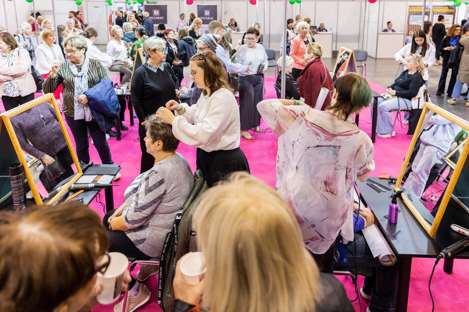 Duży pokój wypełniony ludźmi. Pokój ma różowy dywan i jest ozdobiony zielonymi i białymi balonami. W pokoju jest kilka osób, niektóre stoją, a niektórzy siedzą na krzesłach. W środku jest kobieta o blond włosach, ubrana w białą bluzkę z kwiatowym wzorem. Stoi przed lustrem i wydaje się, że robi włosy. Inni stoją wokół niej, obserwując jej pracę. Niektórzy z nich trzymają kubki kawy, sugerując, że mają przerwę od dnia.