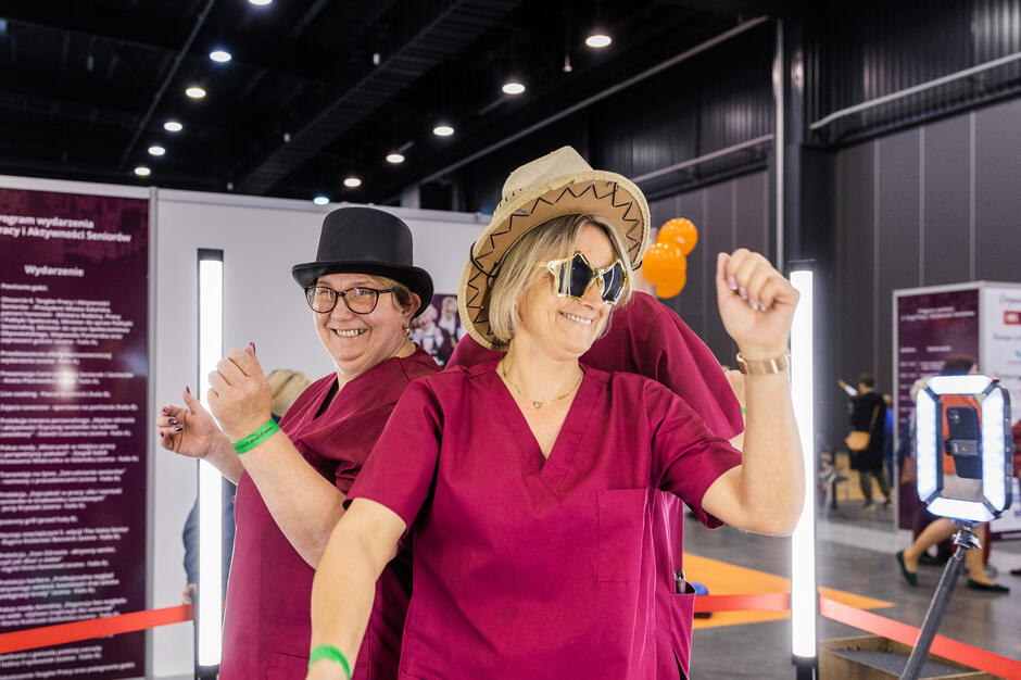 Dwie kobiety w różowych chustach i kapeluszach, stojące w przestrzeni wewnętrznej z czerwoną barierą wstążki w tle. Oboje uśmiechają się i wydają się pozować do zdjęcia. Kobieta po lewej stronie nosi czarny kapelusz i okulary, podczas gdy kobieta z przodu trzyma znak pokoju obiema rękami. Obie kobiety noszą okulary przeciwsłoneczne i mają ramiona podniesione w sposób uroczysty. Na jednej z barier znajduje się napis "Witamy na wystawie" oraz aparat na statywie obok nich. 