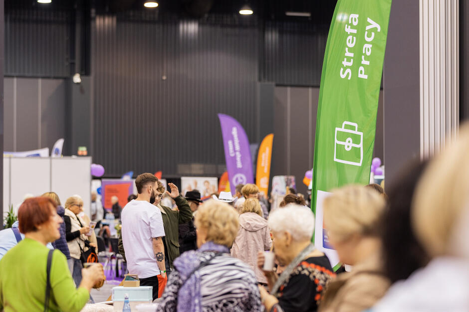 Duża grupa ludzi zebrała się w sali kongresowej, sala ma wysoki sufit z czarnymi panelami i zielonym banerem wiszącym od prawej strony, po lewej stronie stoi przed stołem z laptopem i innymi przedmiotami, nosi białą koszulkę i wydaje się rozmawiać z ludźmi wokół siebie, w tłumie są inni ludzie, niektórzy patrzą na baner, a inni rozmawiają.