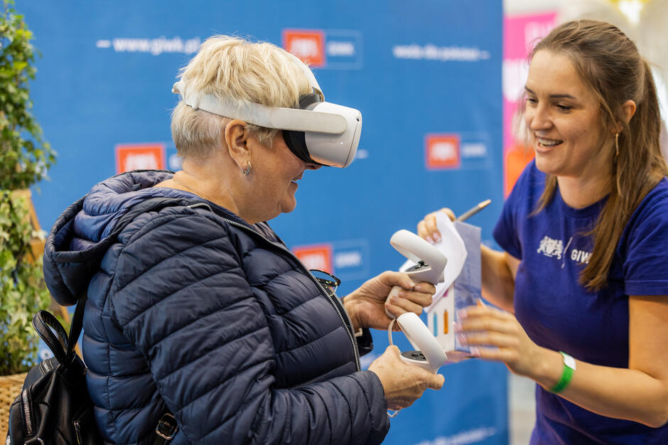 Starsza kobieta w zestawie słuchawkowym wirtualnej rzeczywistości i niebieskiej kurtce stoi przed banerem z logo, trzyma w ręku biały pilot i wydaje się, że współdziała z młodą kobietą, która również ma na sobie zestaw słuchawek, obie kobiety uśmiechają się i zdają się być zaangażowane w rozmowę, wydaje im się, jakby były na wydarzeniu lub wydarzeń mediów społecznościowych.