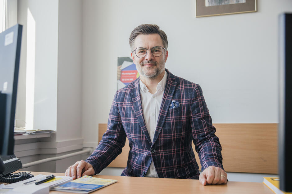 Mężczyzna w średnim wieku, w okularach. Stoi przy biurku, uśmiecha się