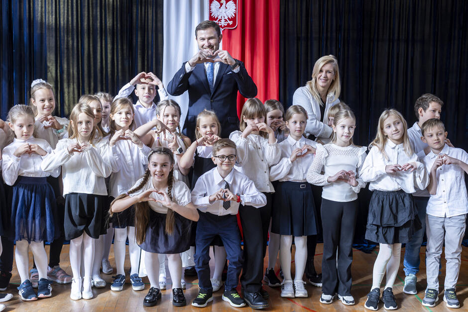 Kobieta i mężczyzna pozują do zdjęcia z grupą odświętnie ubranych dzieci na sali gimnastycznej.