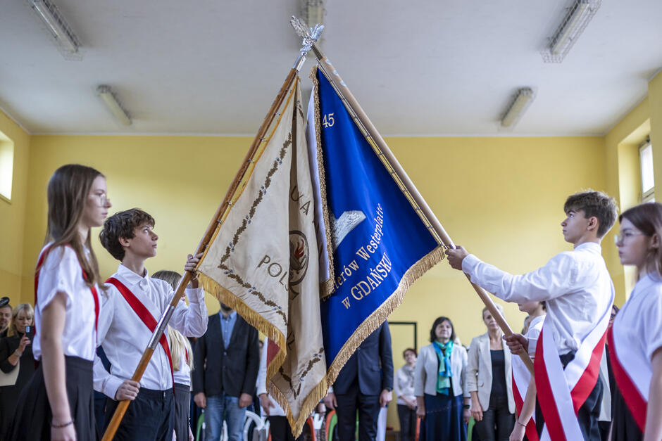 Dwa poczty sztandarowe krzyżują dwa sztandary na sali gimnastycznej w szkole.