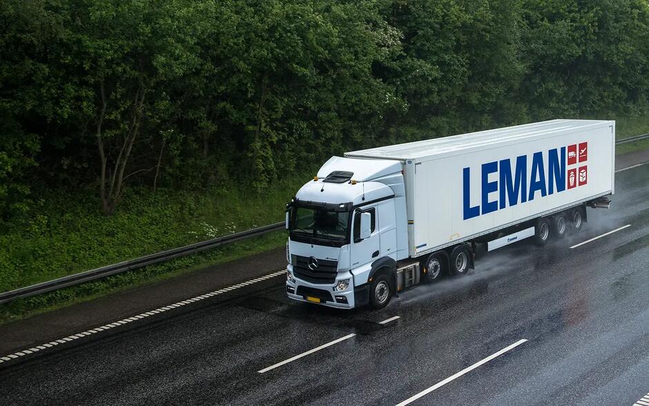 Na zdjęciu widać biały ciągnik siodłowy z naczepą jadący po mokrej autostradzie w deszczowej pogodzie. Na boku naczepy widnieje duży niebieski napis „LEMAN” oraz czerwone piktogramy symbolizujące różne rodzaje transportu i logistyki. Droga jest otoczona gęstym, zielonym lasem, a mokra nawierzchnia odbija światło, tworząc efekt połysku.