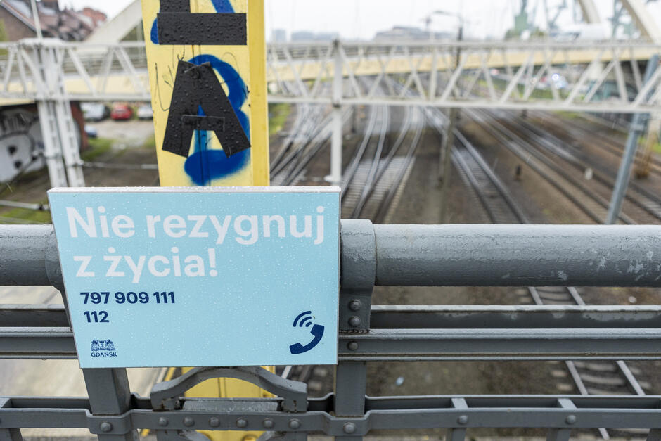 Tabliczka z numerami telefonów i napisem Nie rezygnuj z życia  zawieszona na barierce mostu.