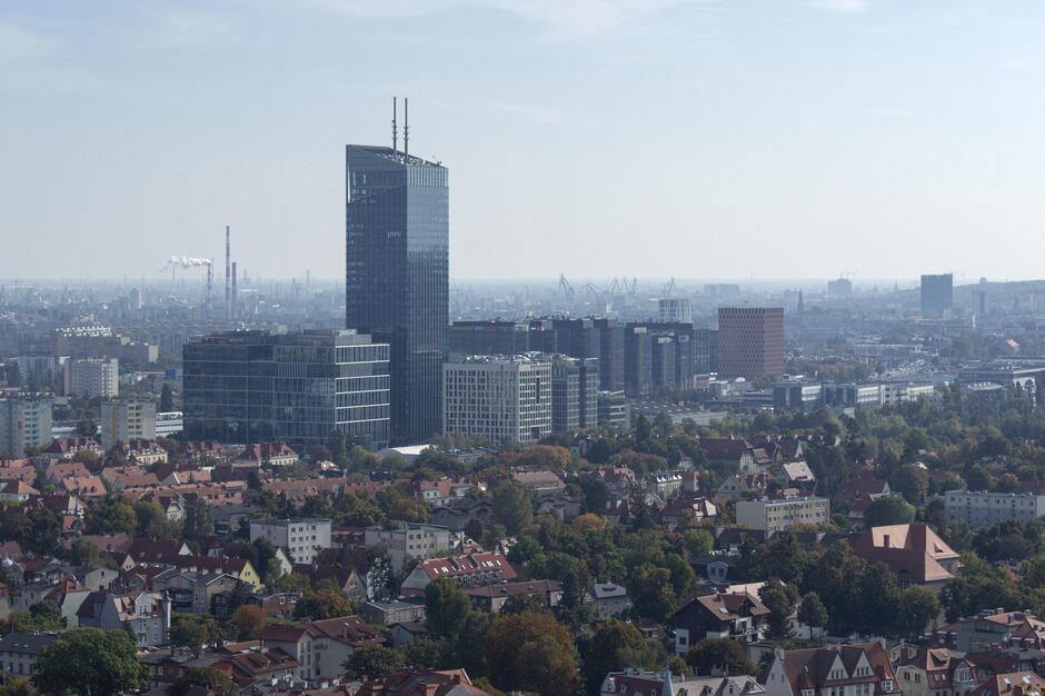 Panorama miasta z widocznym nowoczesnym kompleksem biurowców, wśród których dominuje wysoki szklany wieżowiec. Na pierwszym planie rozciąga się zabudowa z czerwonymi dachami, otoczona drzewami i zielenią miejską.
