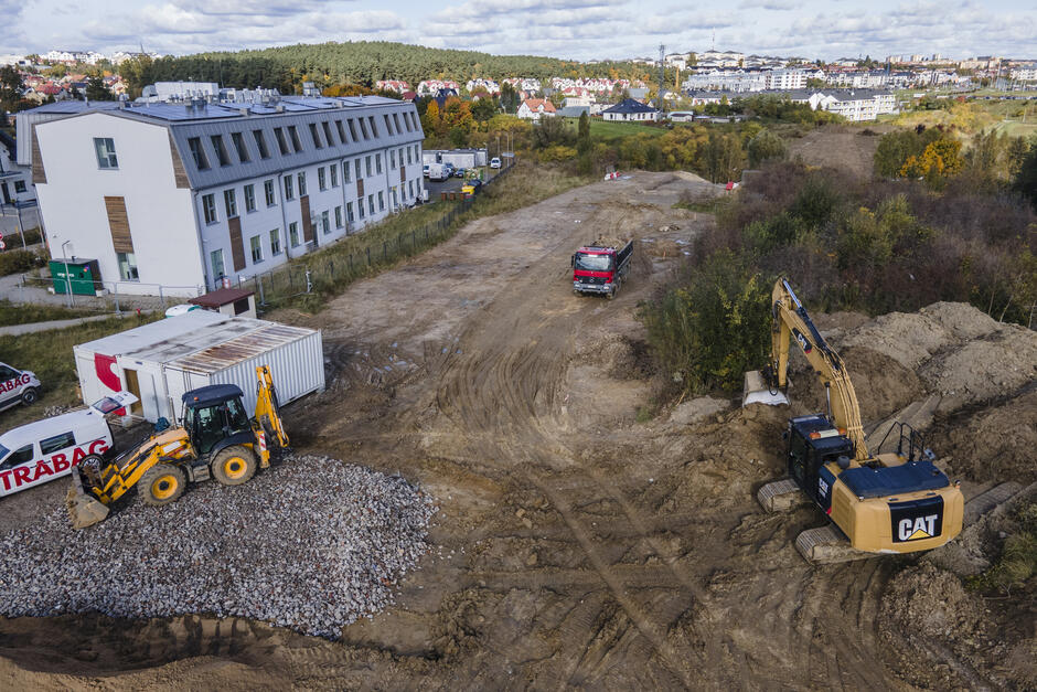 na zdjęciu plac budowy widać żółte koparki i dwie ciężarówki, do tego rozkopany teren gruntowy, w tle biały budynek