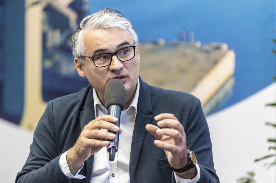 Mężczyzna w okularach i marynarce mówi do mikrofonu podczas dyskusji, gestykulując dłonią. W tle widoczna jest rozmyta grafika przedstawiająca fragment nadmorskiego krajobrazu. 