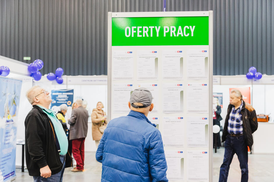 na zdjęciu zielony kubik na którym wiszą ofert pracy, czyta je trzech seniorów