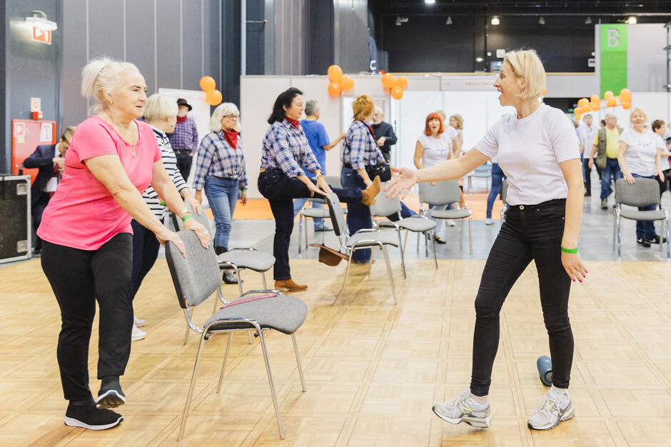 na zdjęciu kilka starszych ćwiczących kobiet, naprzeciwko jednej z nich stoi kobieta w średnim wieku, coś jej tłumaczy