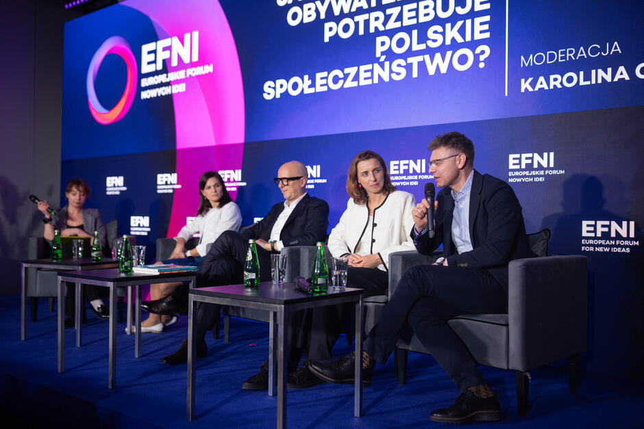 Grupa ludzi w eleganckich strojach siedzi na scenie, za nimi duży ekran z napisem EFNI  